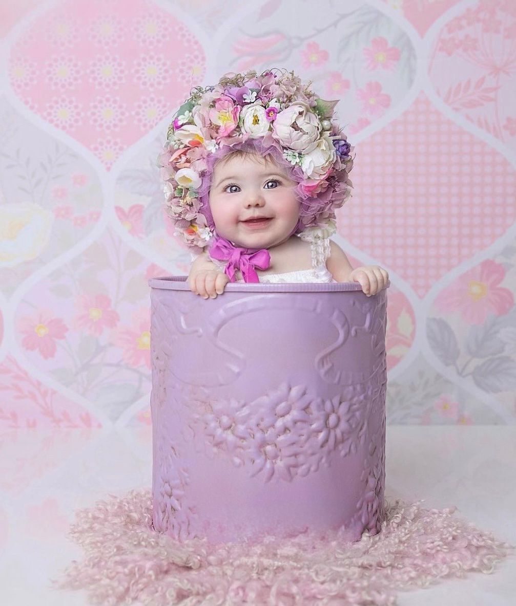 Adorable baby sitting in purple decorative cylinder wearing floral crown against pink heart background.