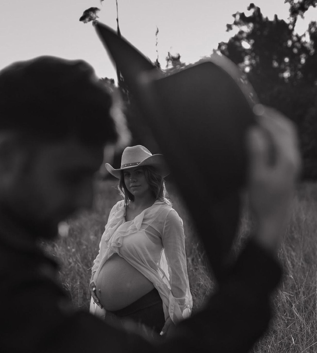 Black and white maternity photo of pregnant woman in cowboy hat and white dress standing in a field.