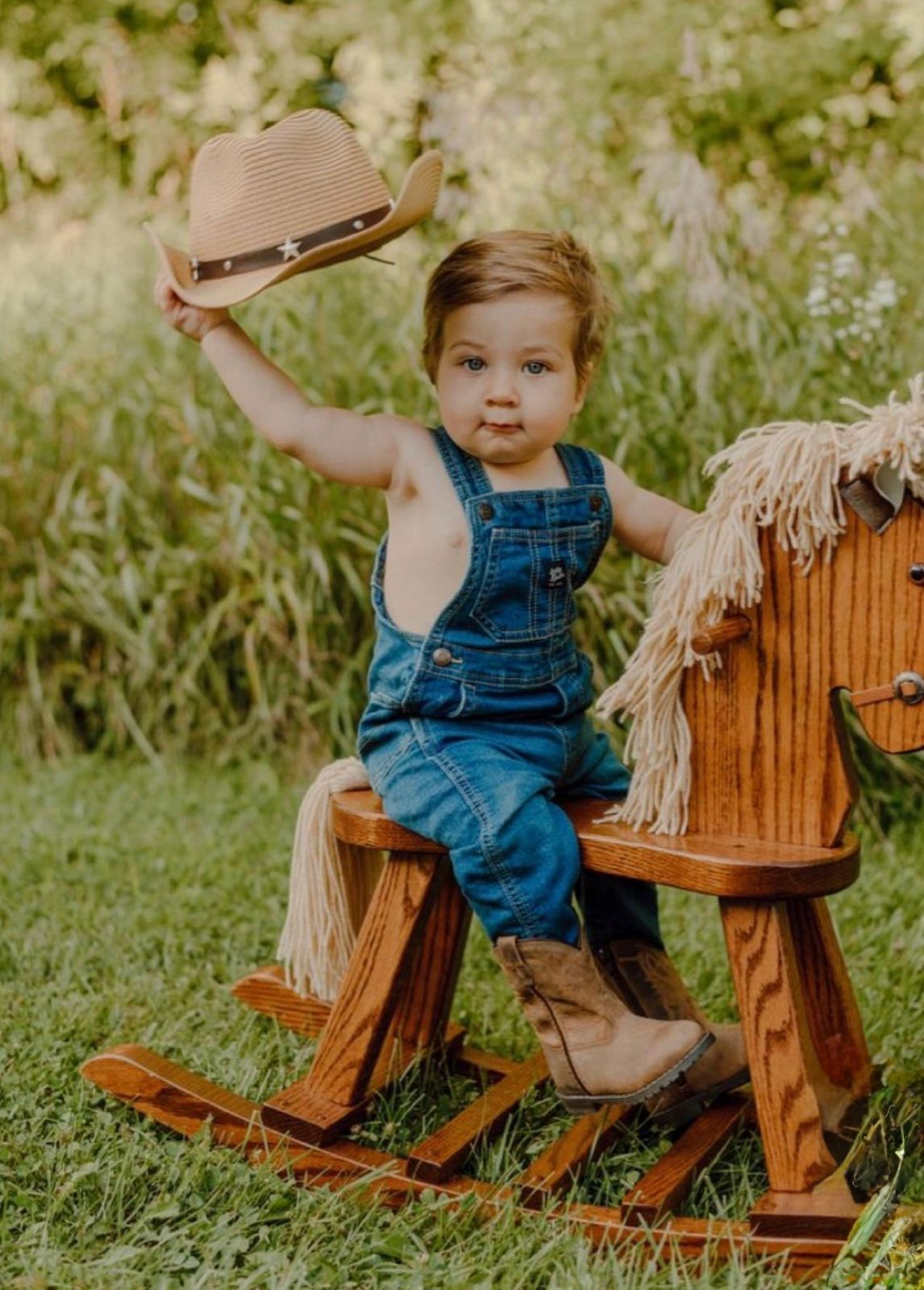 Toddler boy in denim overalls and cowboy boots sits on wooden rocking horse, tipping straw cowboy hat outdoors.