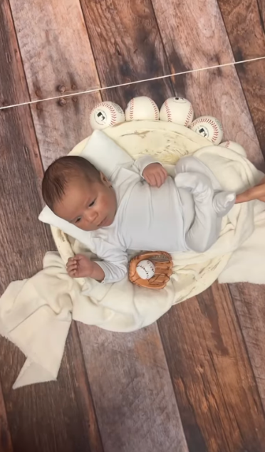Baby in white sleeper lying on cream blanket with baseballs on wooden floor backdrop.