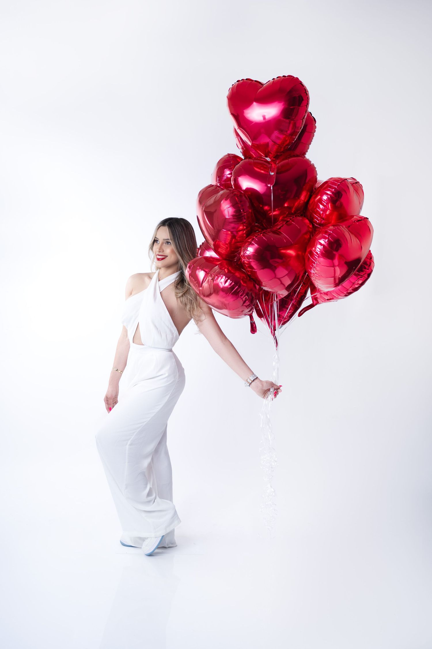 Elegant figure in white dress poses with cluster of red heart balloons in studio setting.