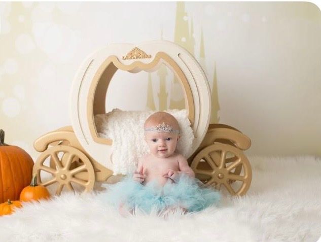 Baby girl in blue tutu sits in wooden pumpkin carriage prop with pumpkins and white fur backdrop.