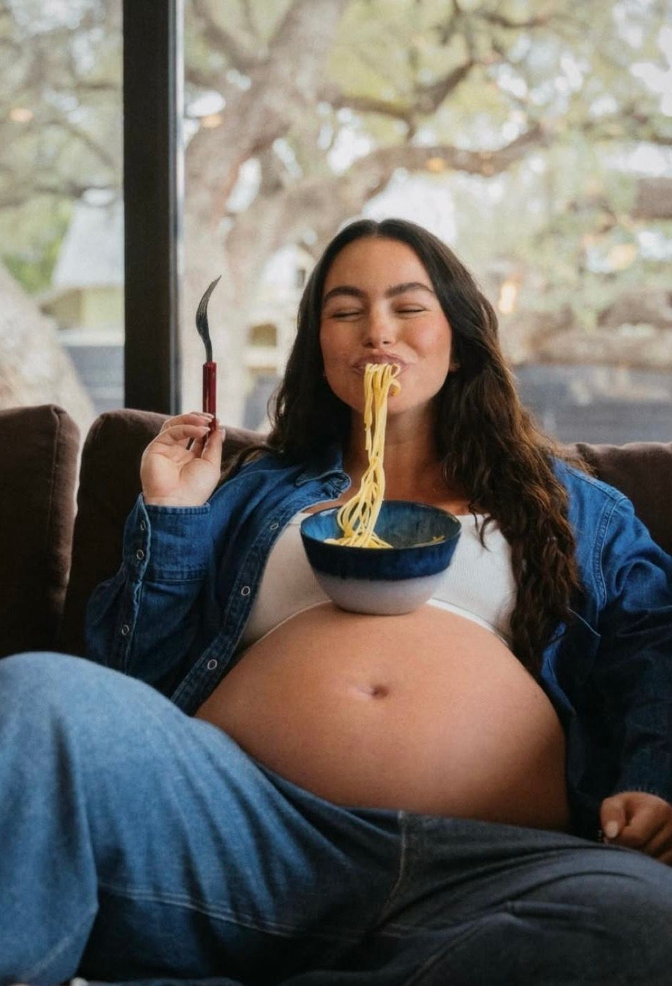 Pregnant woman joyfully eating noodles from a blue bowl while relaxing on a brown couch indoors.