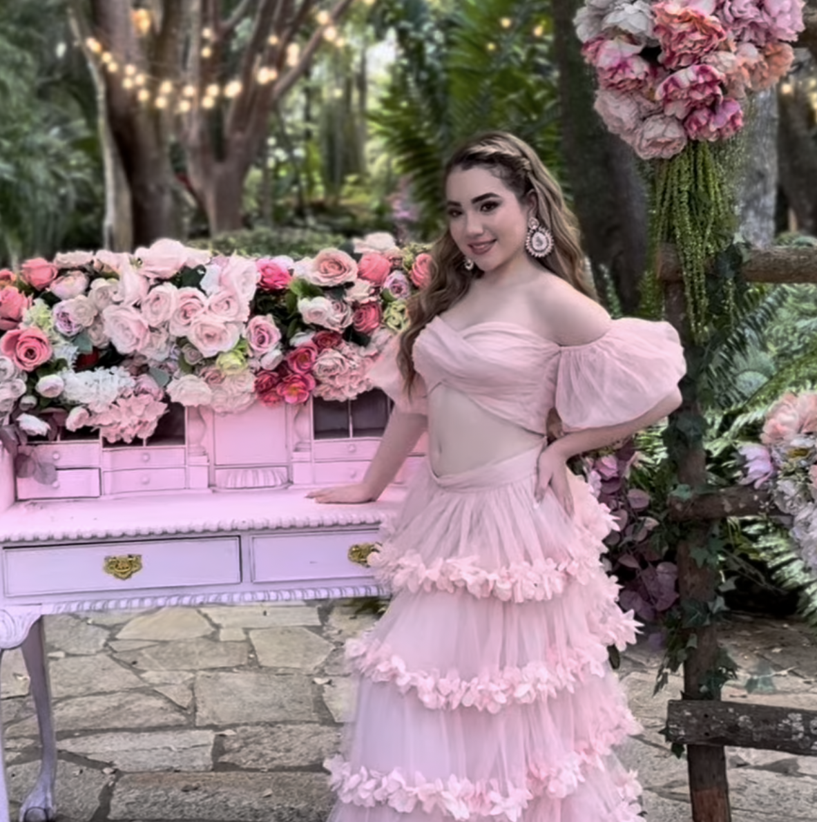 Young woman in pink off-shoulder ruffled gown poses near pink piano with floral arrangements outdoors.