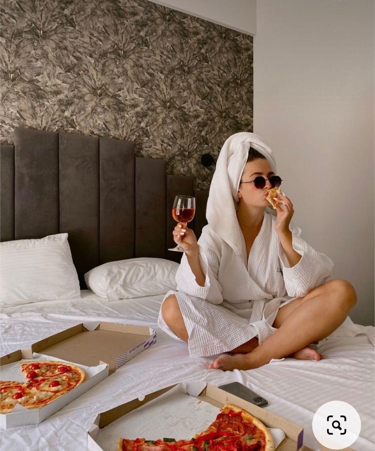 Woman in white robe and towel turban eating pizza and drinking wine on a hotel bed.
