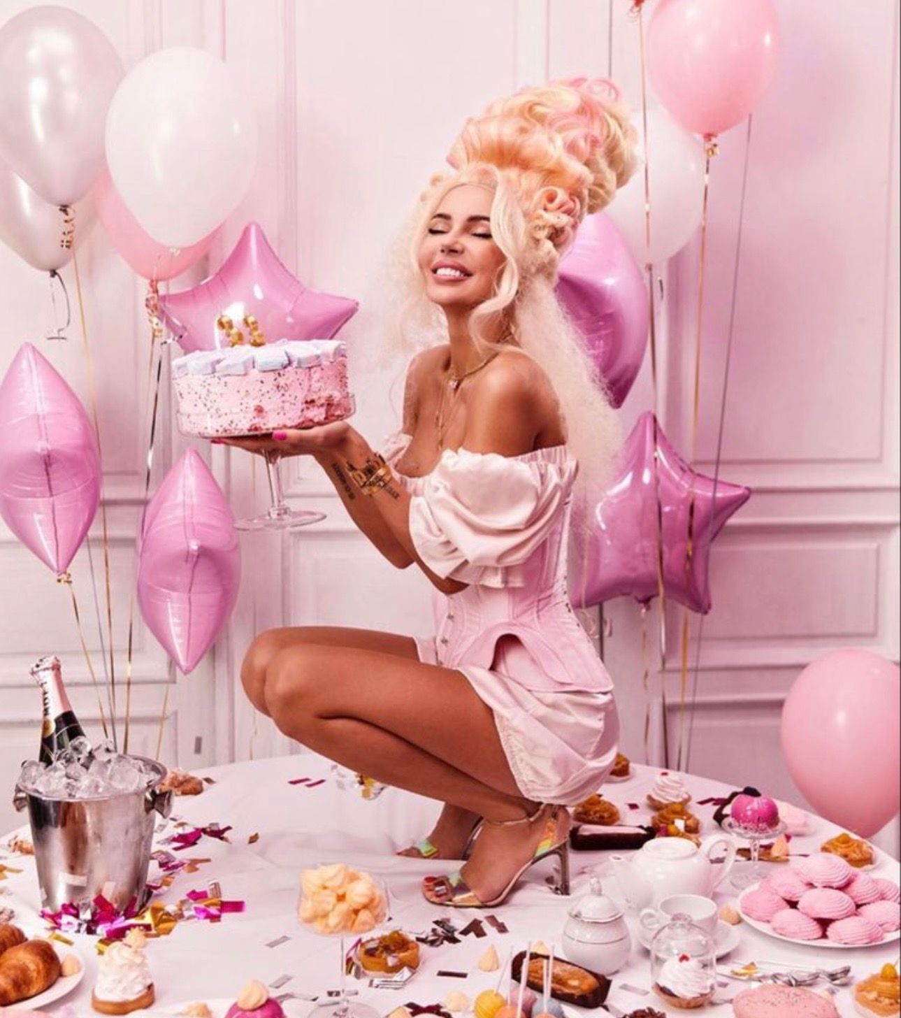 Blonde woman in pink dress holding birthday cake surrounded by balloons, confetti, and sweets in a pink party setting.