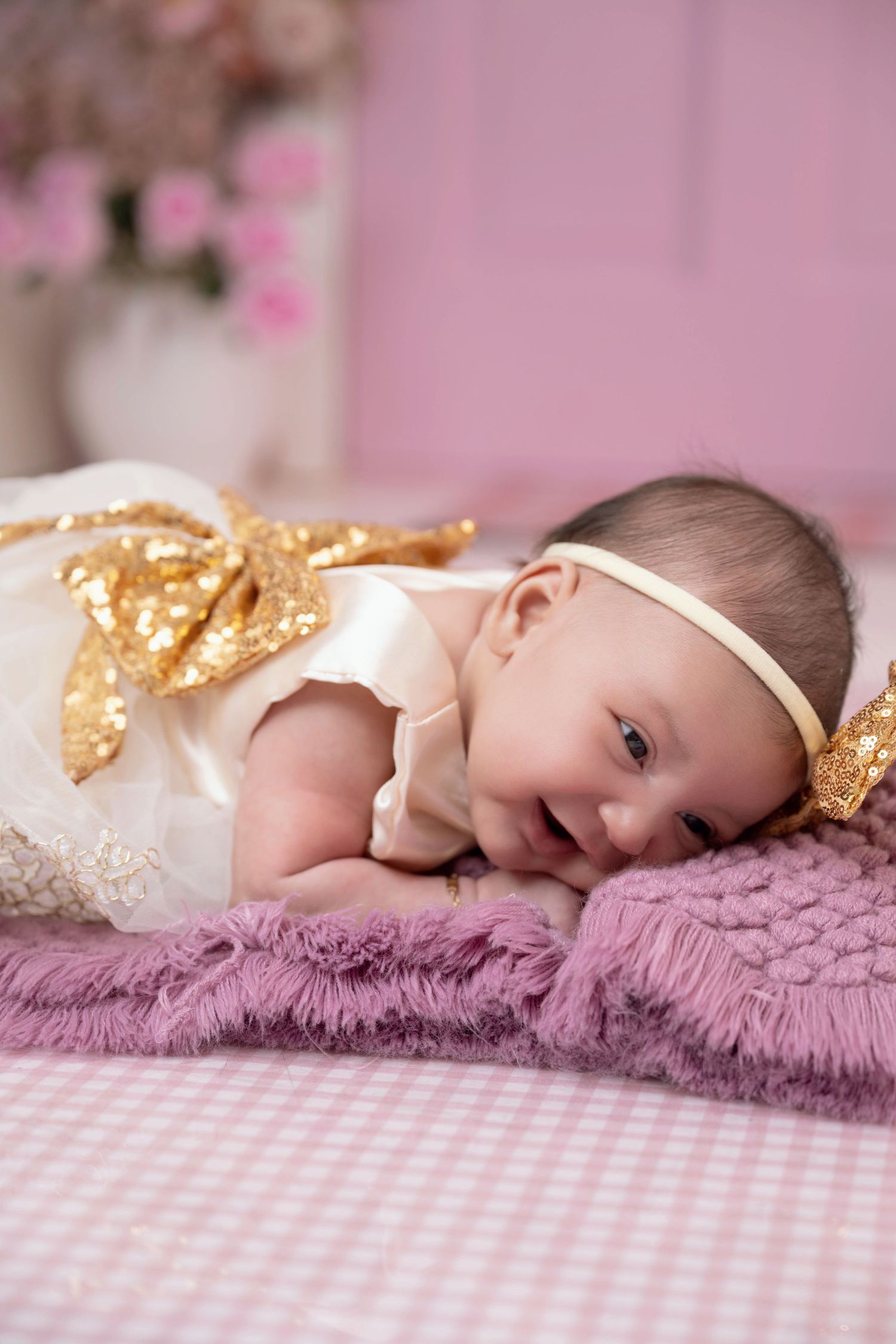 Newborn baby girl in white and gold sequin bow dress with headband lying on purple fuzzy blanket in pink room.