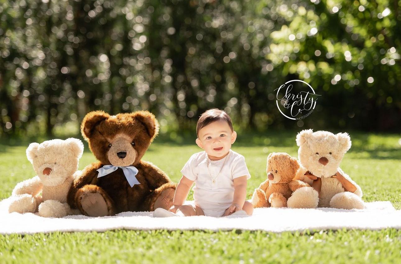 Baby boy sitting on white blanket outdoors surrounded by four teddy bears on green grass.