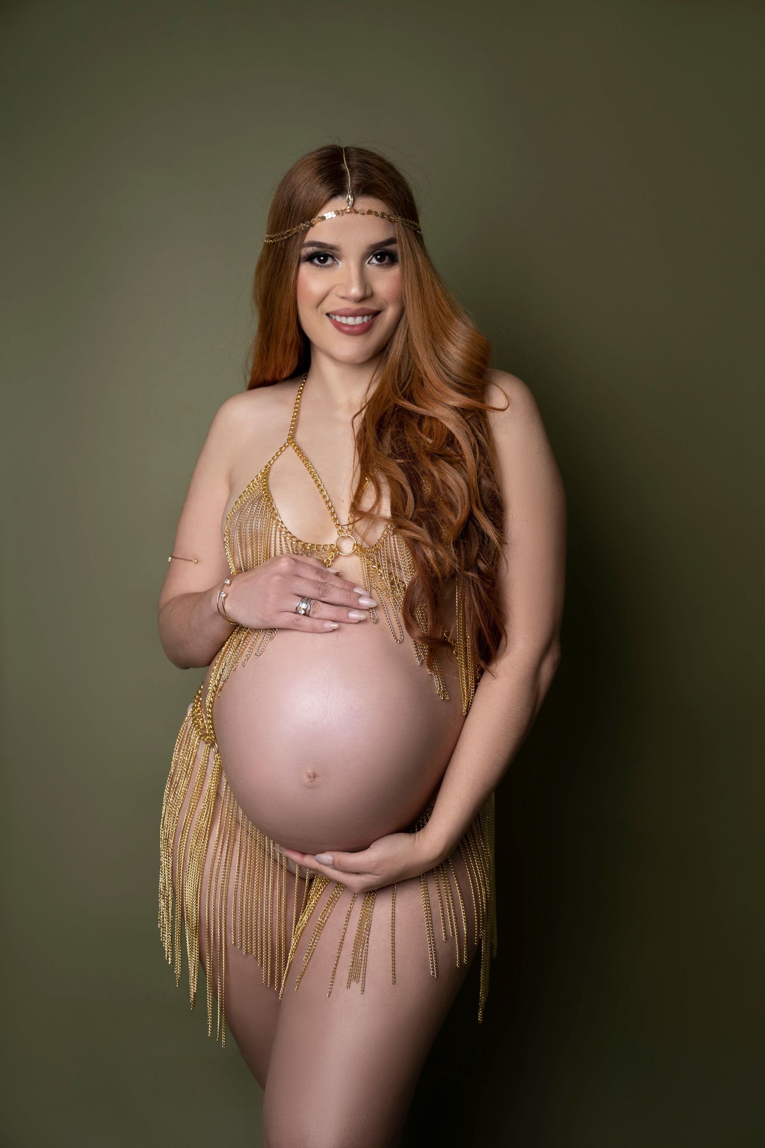 Artistic maternity photo featuring a nude pregnant figure wearing a golden fringe dress against a dark green background.