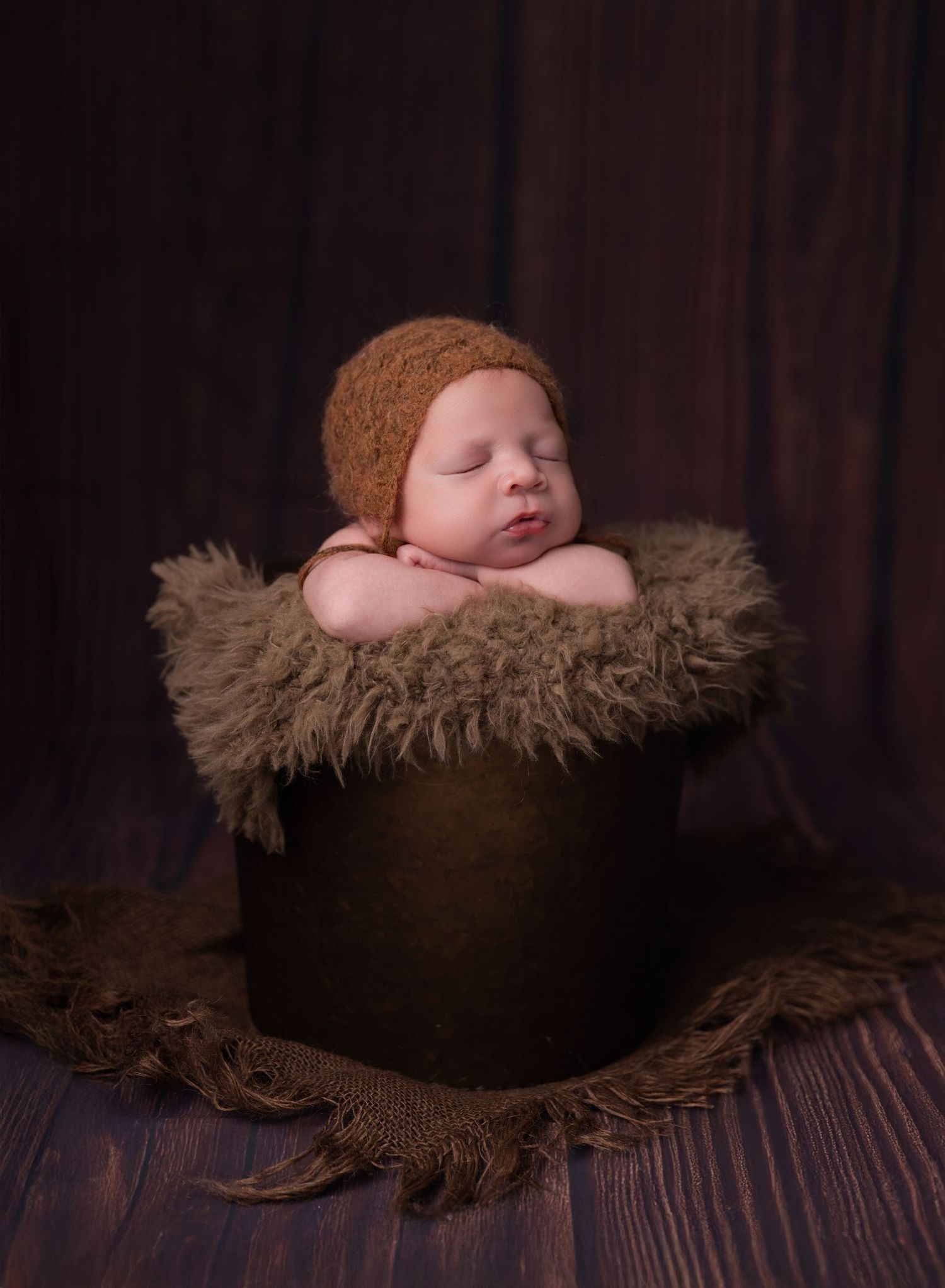 Sleeping newborn baby in brown knit bonnet nestled in a dark bucket with fluffy fur, studio portrait photography.