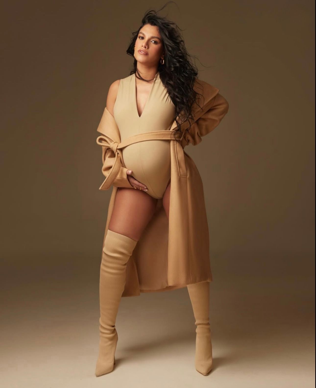 Pregnant woman poses in monochromatic camel outfit with wrap coat, bodysuit, and thigh-high boots against warm brown backdrop.