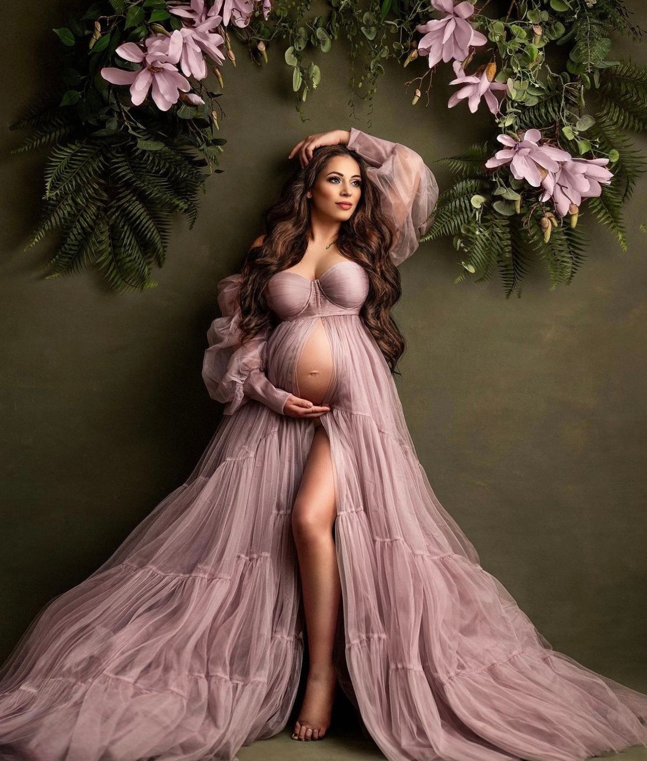 Pregnant woman in flowing mauve tulle gown poses gracefully under floral arch with pink magnolias and ferns.