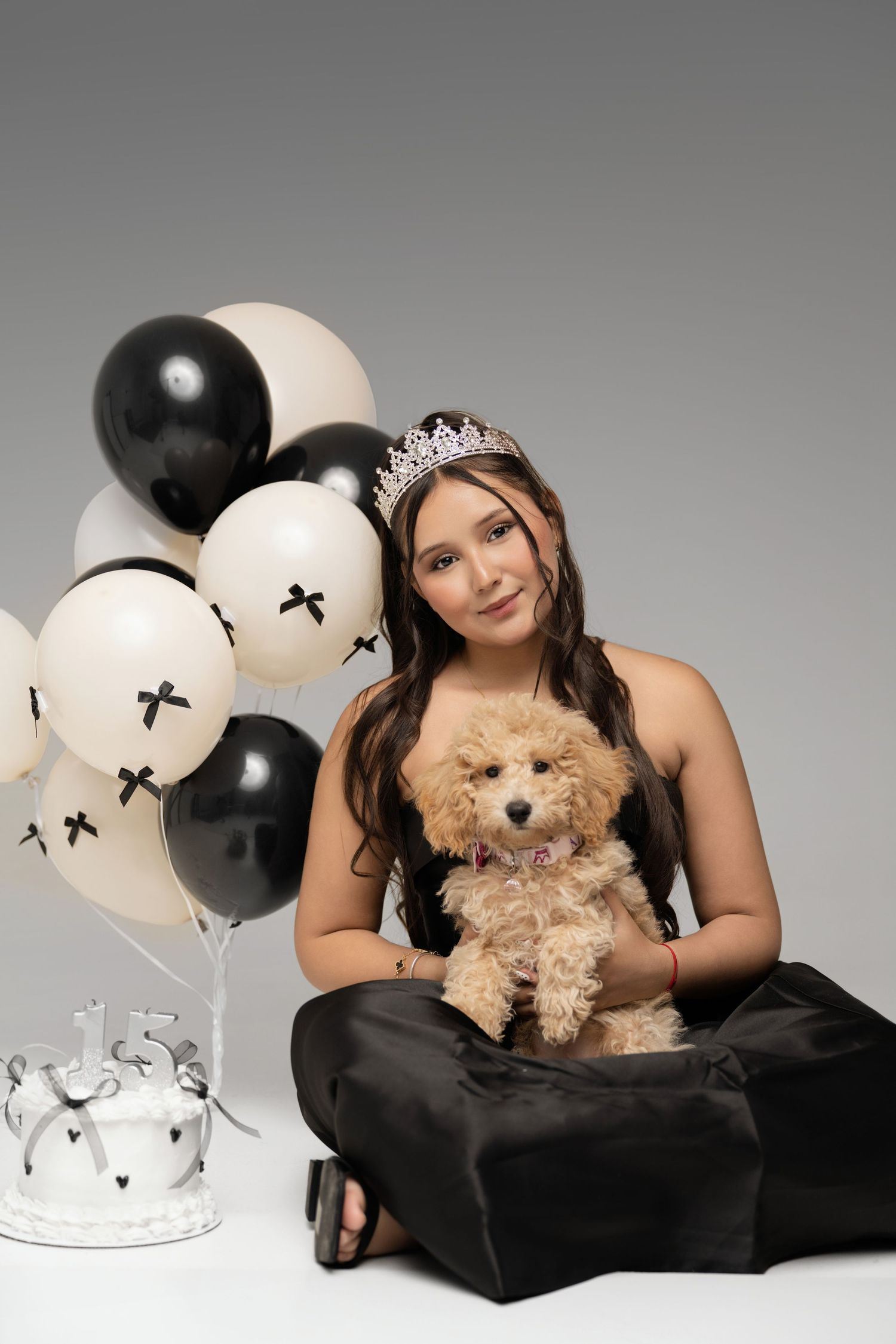 Teen girl in black dress and tiara holds fluffy puppy beside birthday cake and black and white balloons.