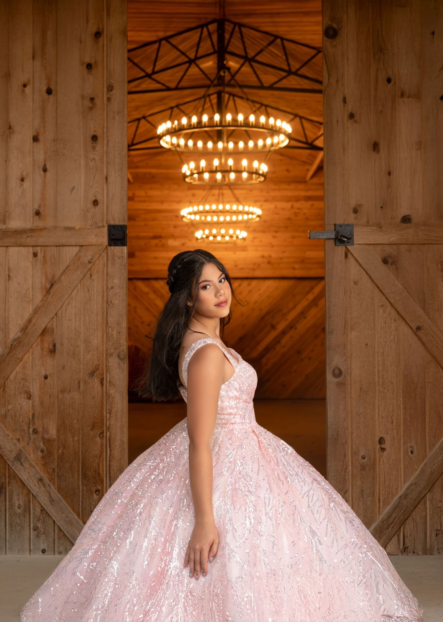 Young woman in a pink sequin ball gown poses before rustic barn doors under warm chandelier lighting.