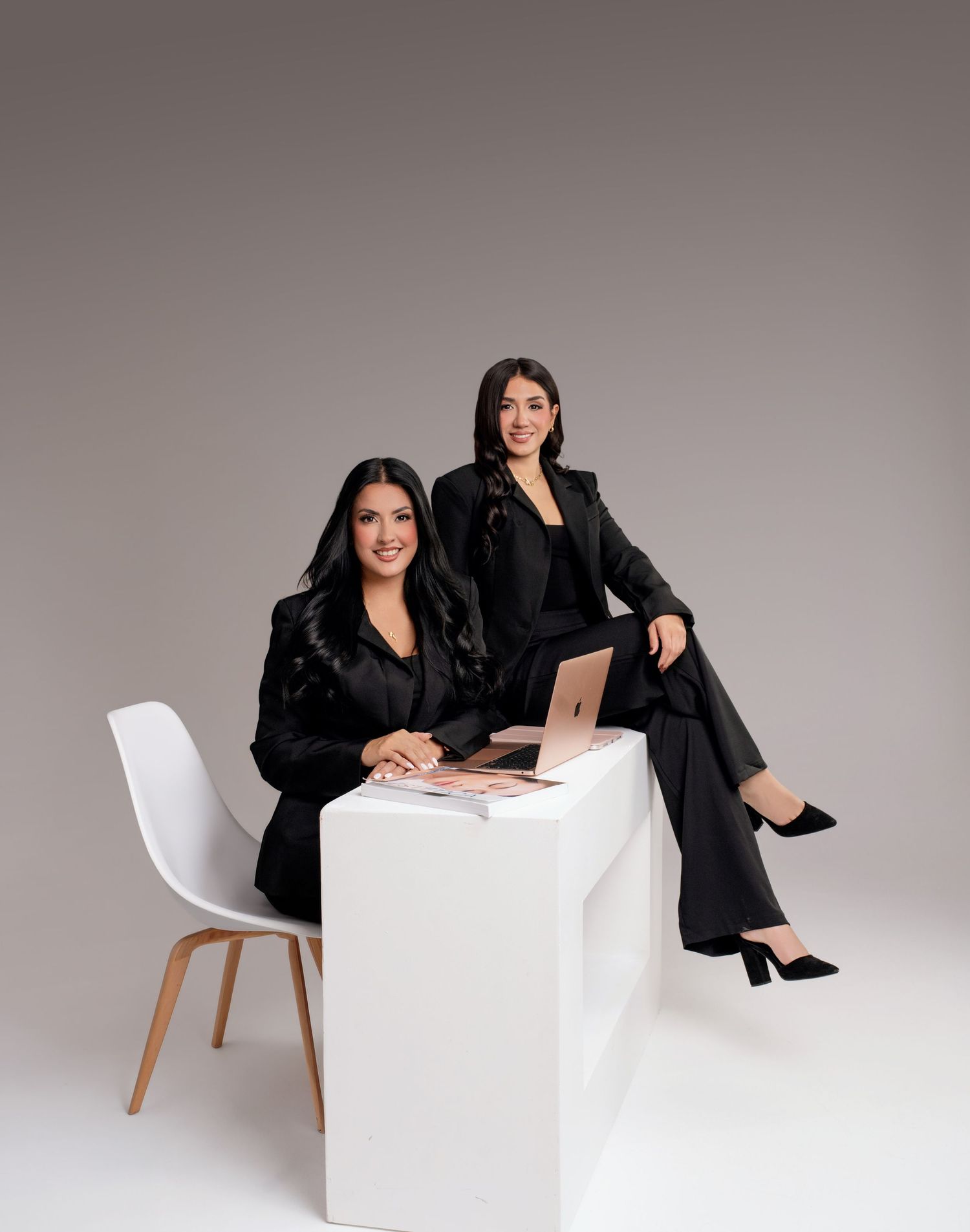 Two professional women in black suits pose confidently with a laptop on a white cube in a modern studio setting.