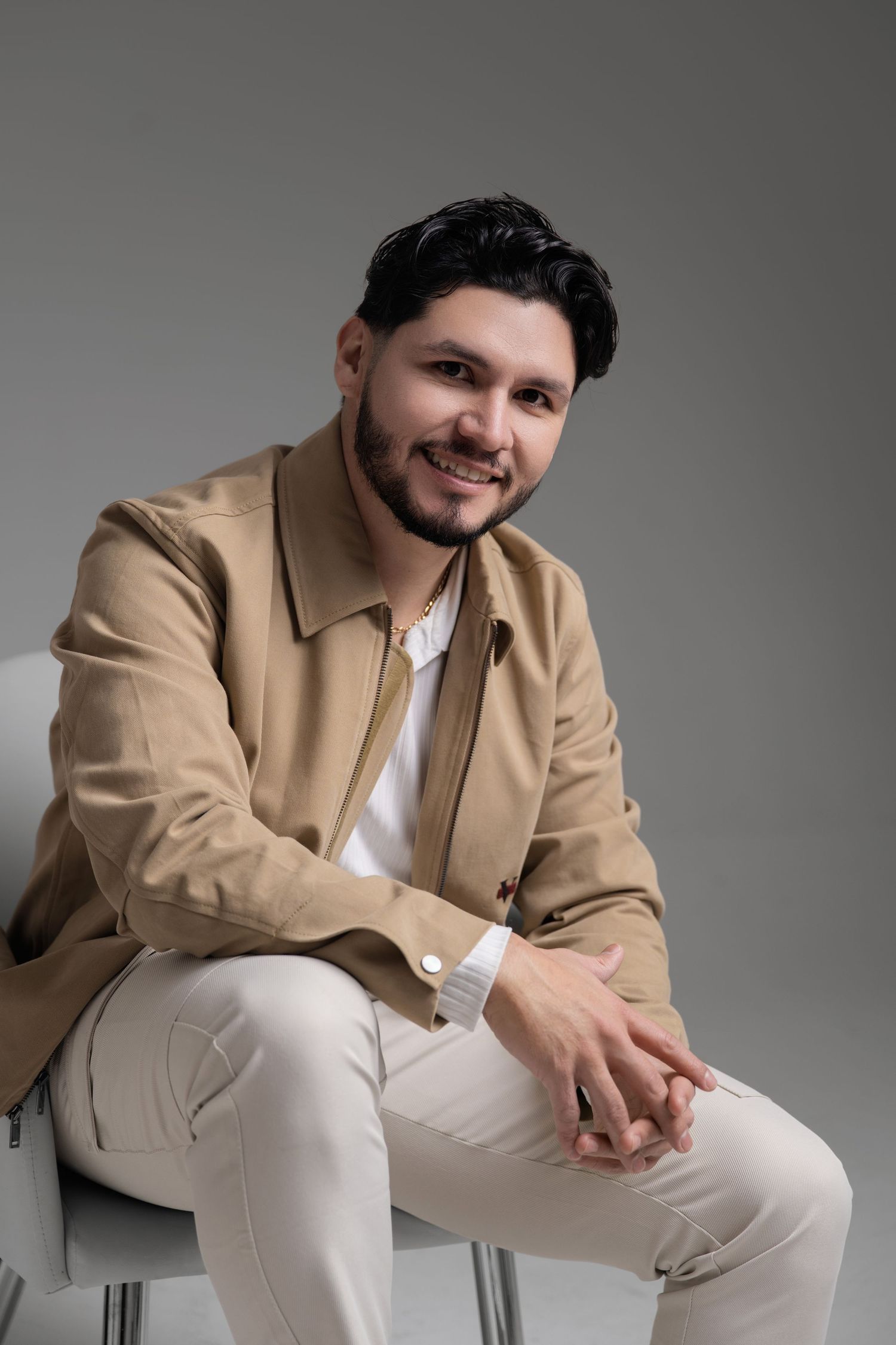 Smiling man with beard wearing beige jacket and white pants sitting relaxed on chair against gray background.