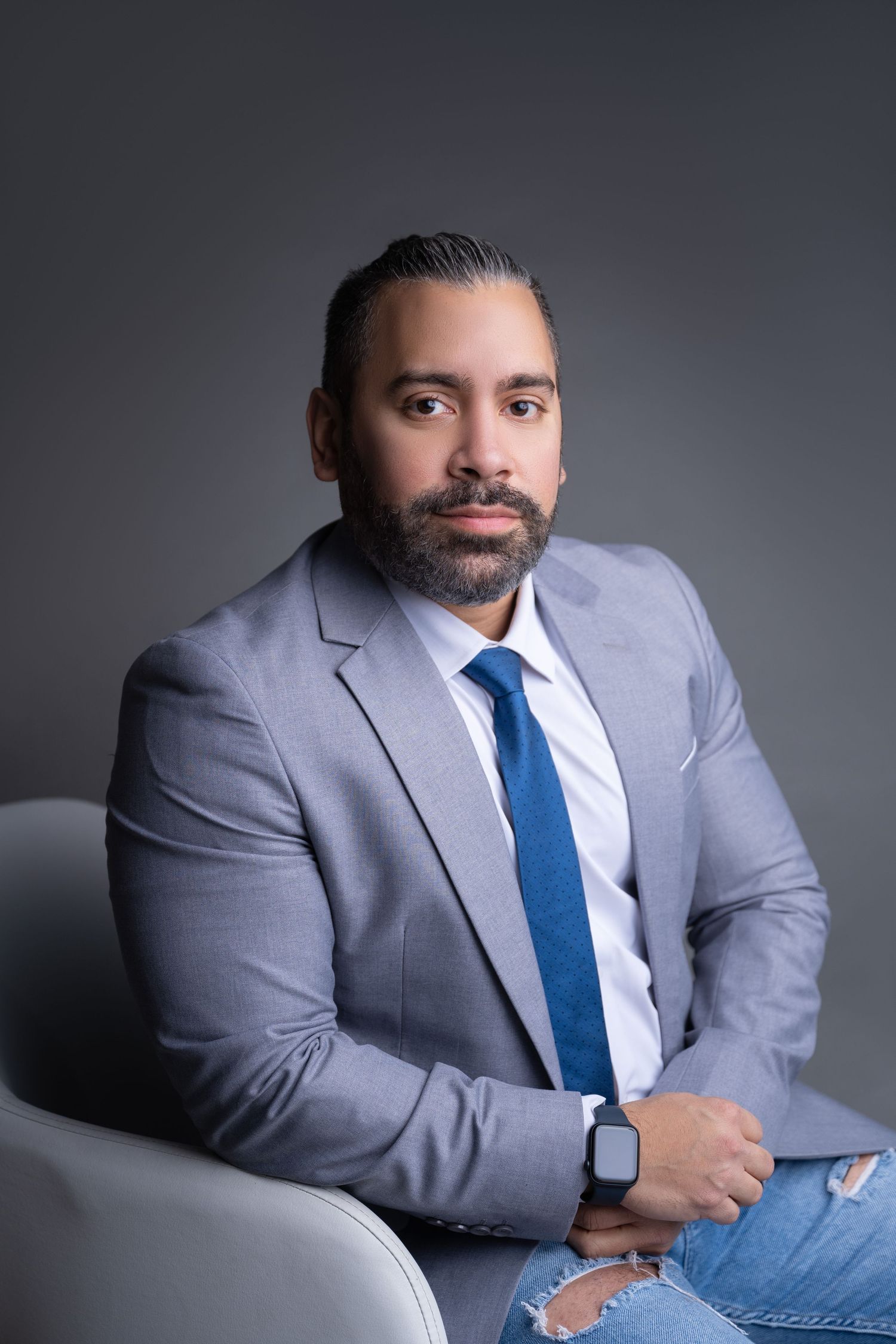 Professional man in gray blazer and blue tie seated confidently, wearing a smartwatch, against a gray studio background.
