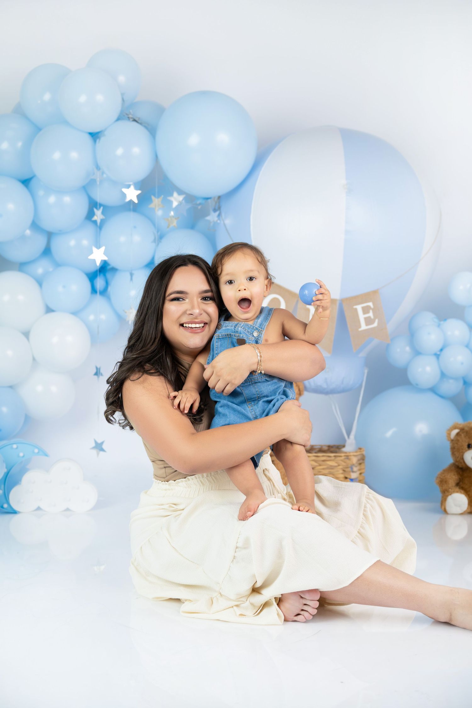 A smiling mother holds her happy baby girl in a blue balloon-themed photo studio setting.