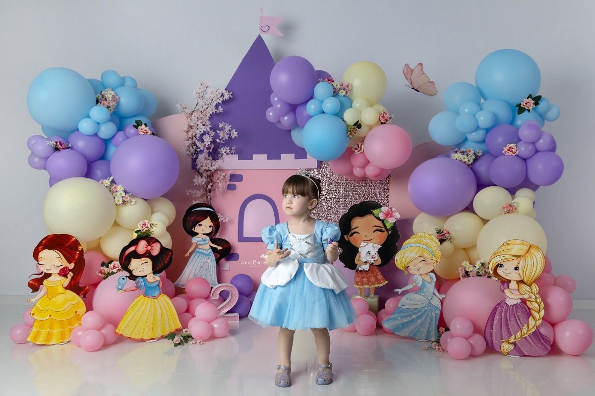 A young girl in a blue Cinderella dress poses at a Disney Princess themed birthday party with colorful balloons and character cutouts.