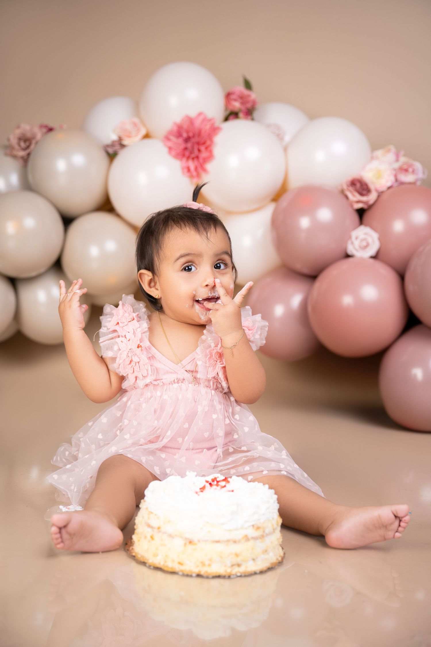 Cute baby girl in pink dress smashing her first birthday cake, surrounded by pink and white balloons.