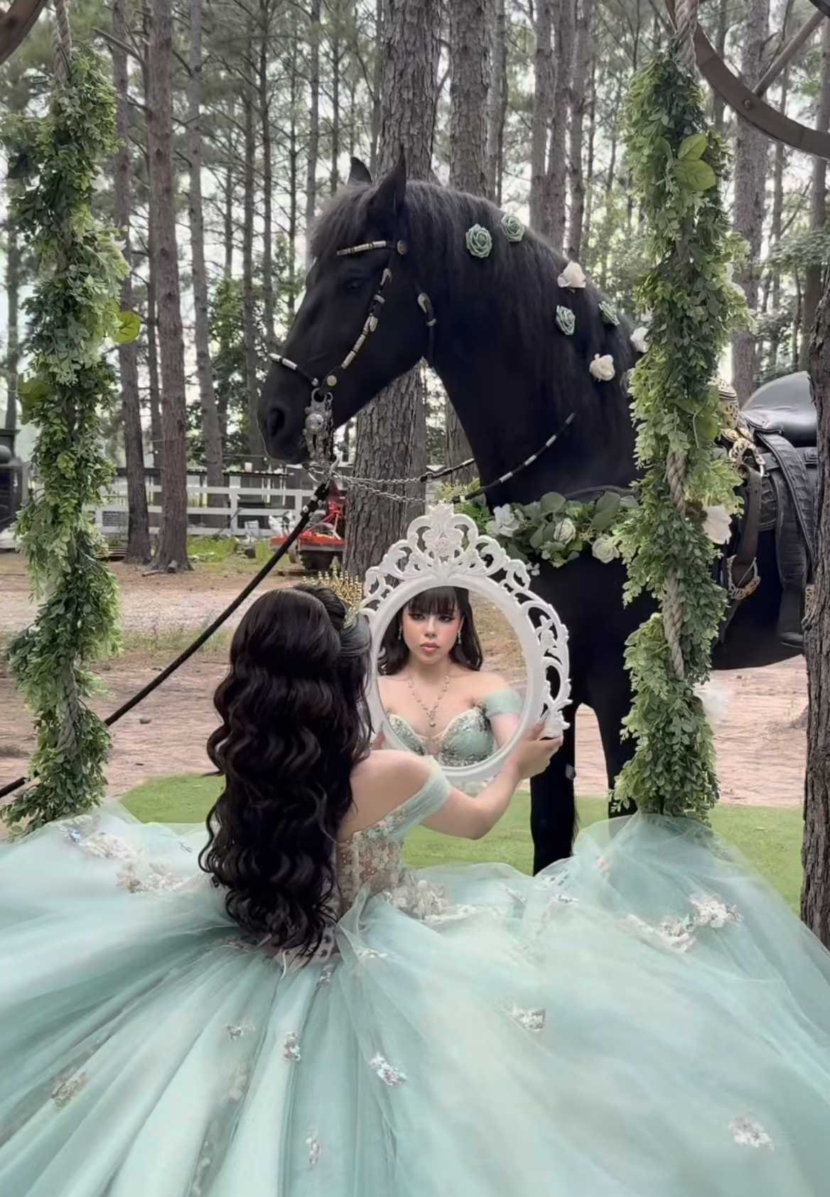 Young woman in mint green quinceañera gown holds ornate mirror beside decorated black horse in forest.