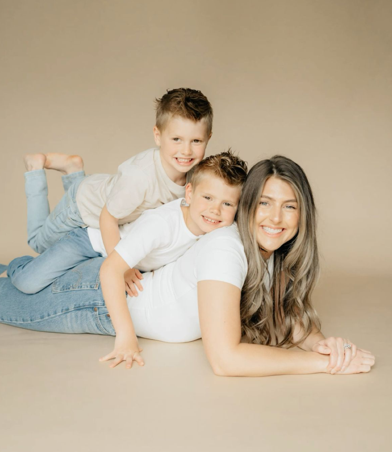 A smiling woman lies on the floor with two young boys stacked on her back in a studio portrait.