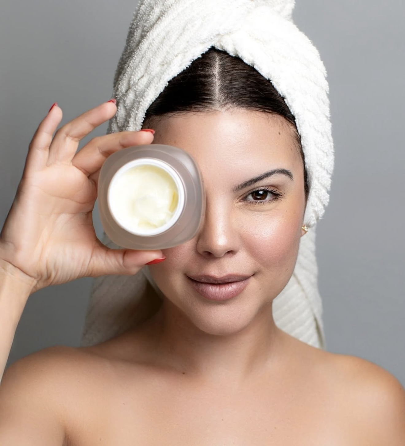 Woman with towel on head holding open jar of face cream over her eye, showcasing skincare moisturizer product.