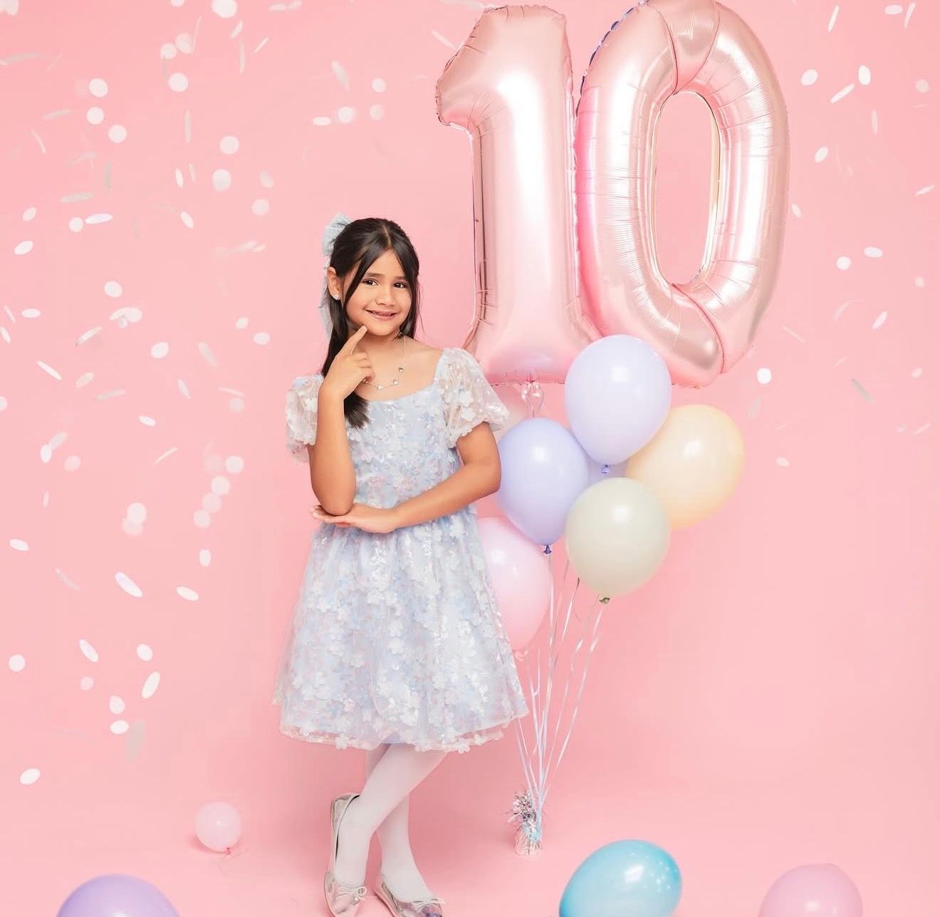 A young girl in a floral dress poses with pastel balloons and large pink number 10 balloons on a pink background.