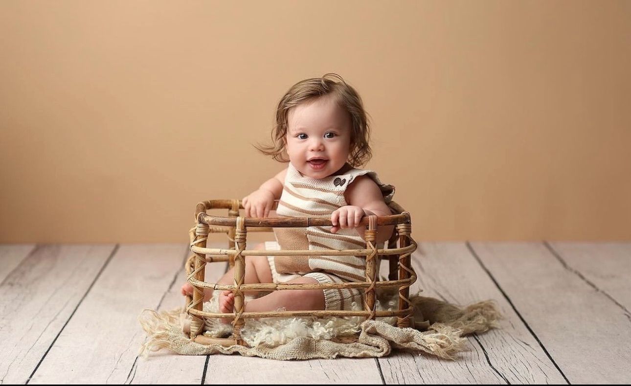 A baby sits inside a rustic wooden crate on a light wooden floor against a neutral background.