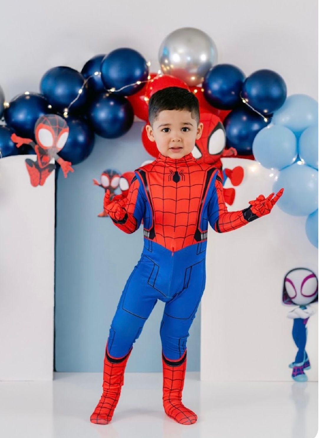 A small child in a Spider-Man costume poses playfully against a backdrop of blue and red balloon decorations.