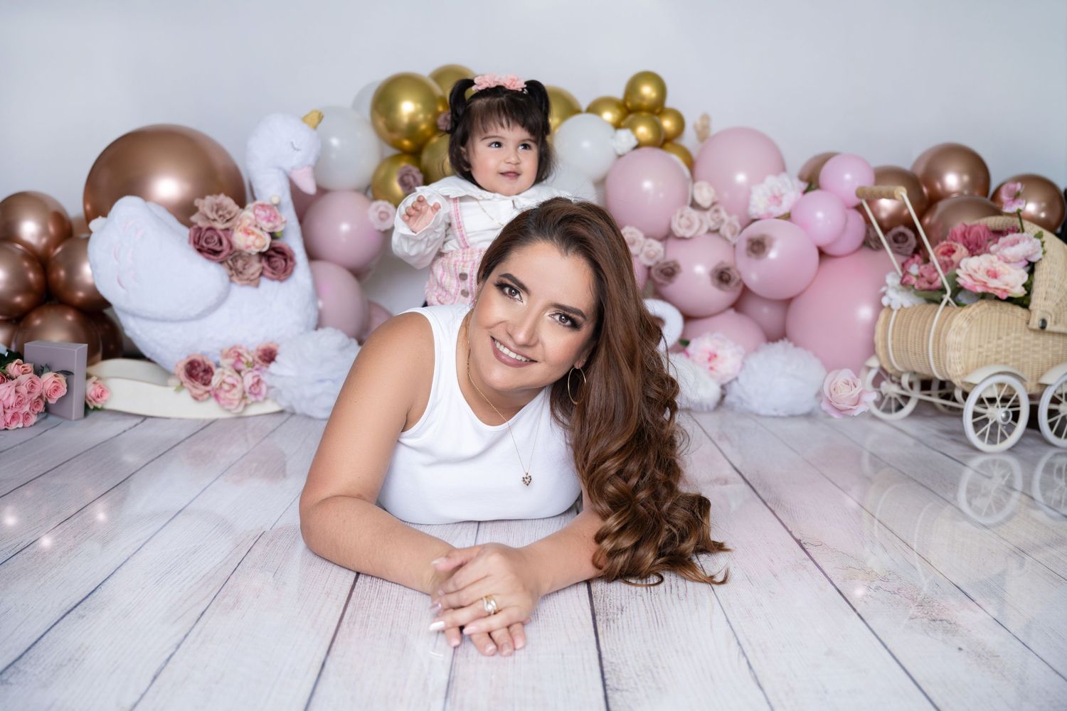 Woman lying on floor smiling with baby girl sitting among pink, gold balloons and floral decorations.