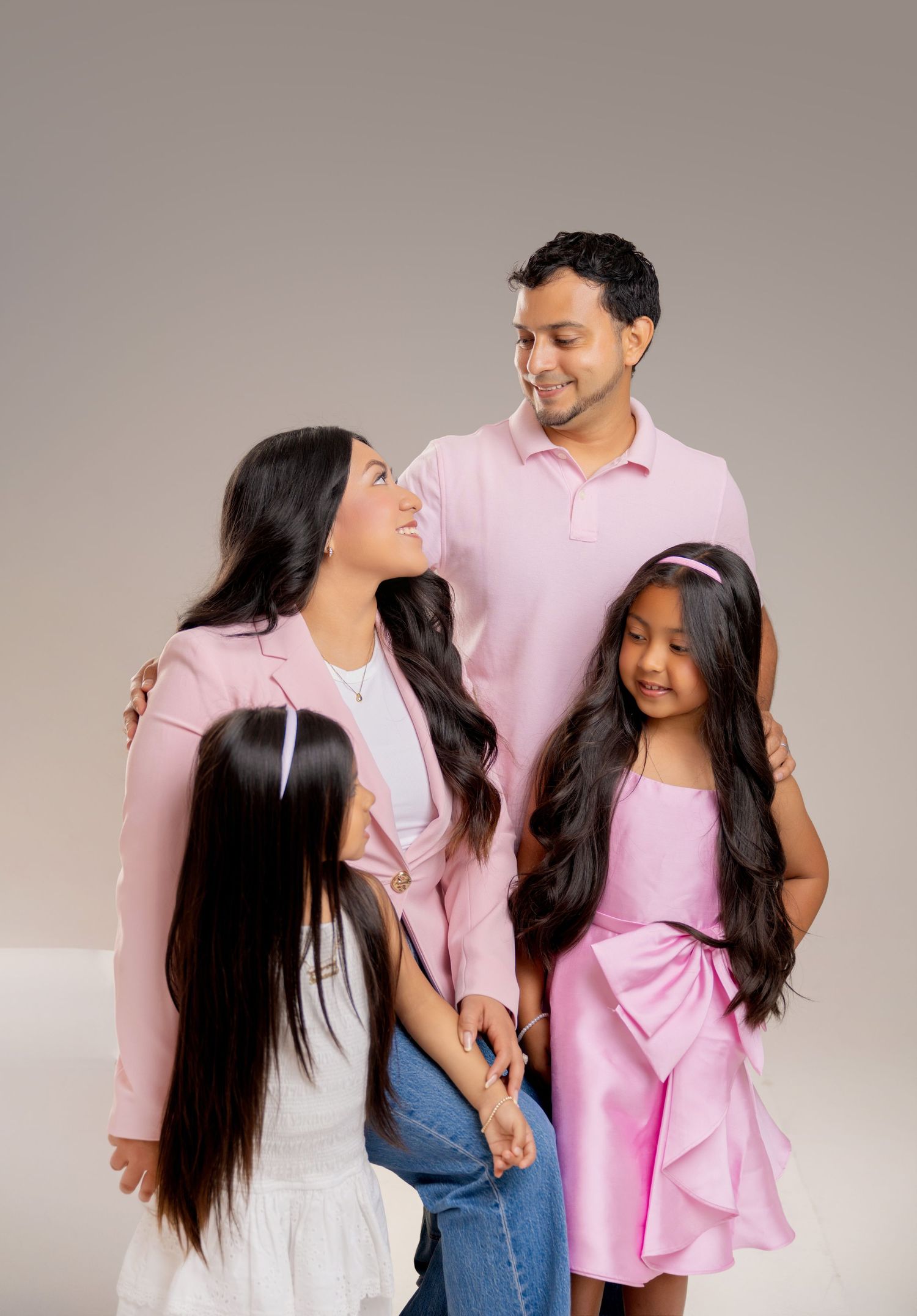 A happy South Asian family of four posing together in coordinating pink outfits against a neutral gray background.