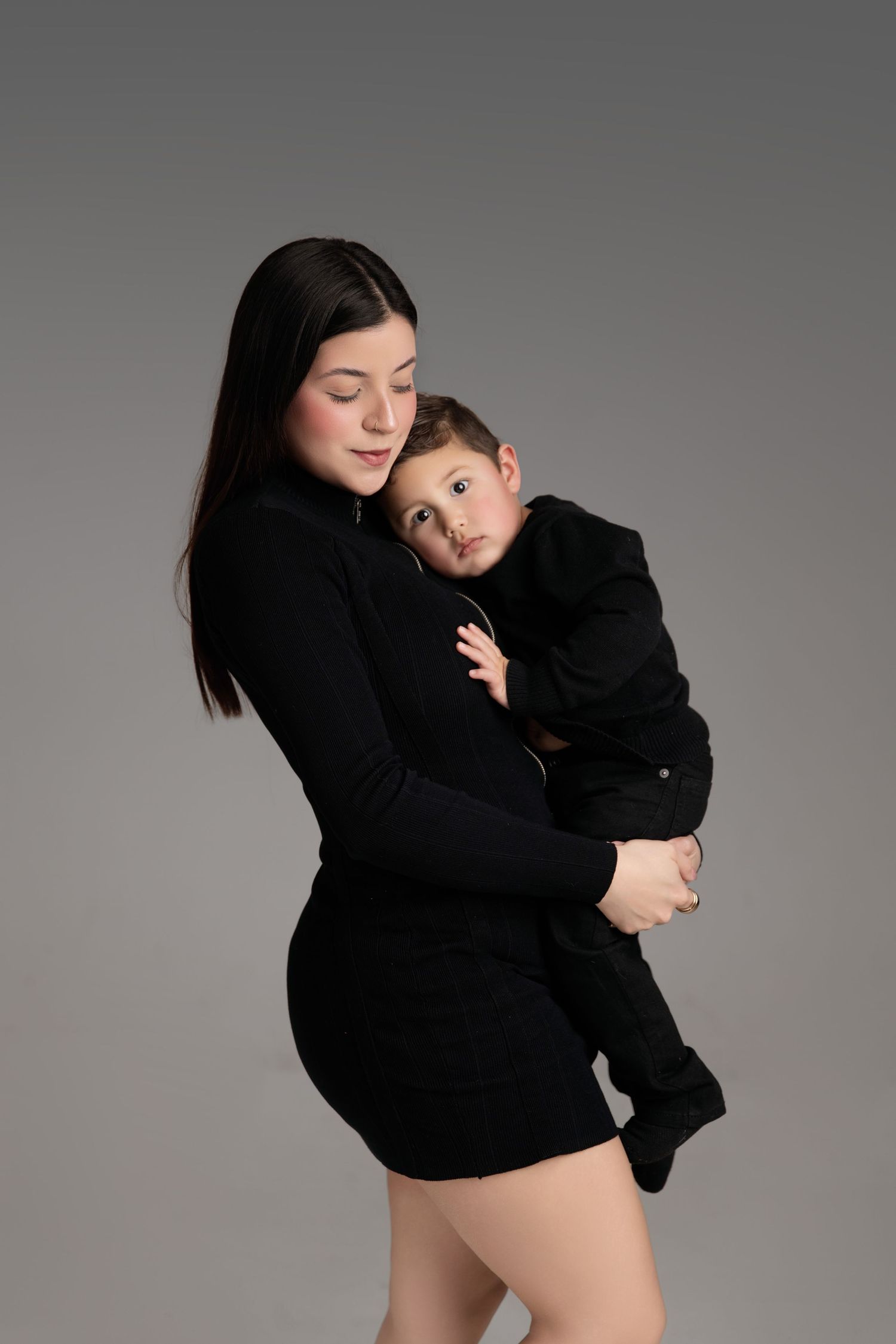 A young mother in a black dress tenderly holds her baby boy, both dressed in black, against a gray studio background.