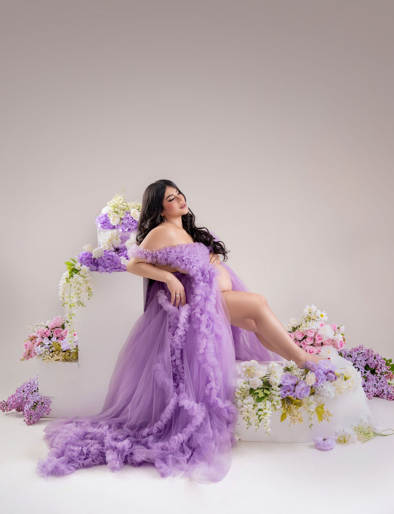 Pregnant woman in lavender ruffle tulle gown poses gracefully among colorful spring flowers in a soft studio setting.