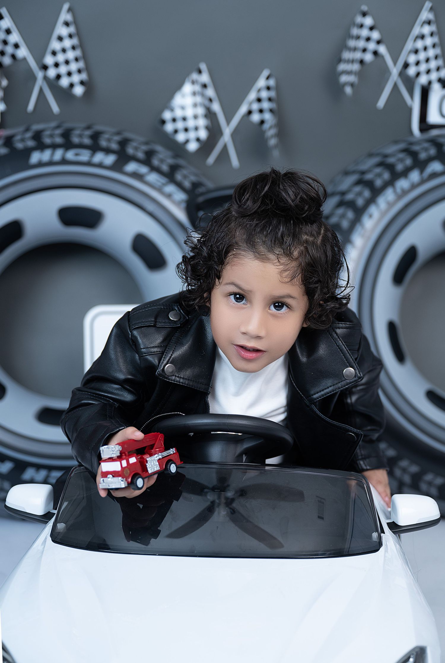 Young child in leather jacket sits in toy white car holding red fire truck with racing flags backdrop.