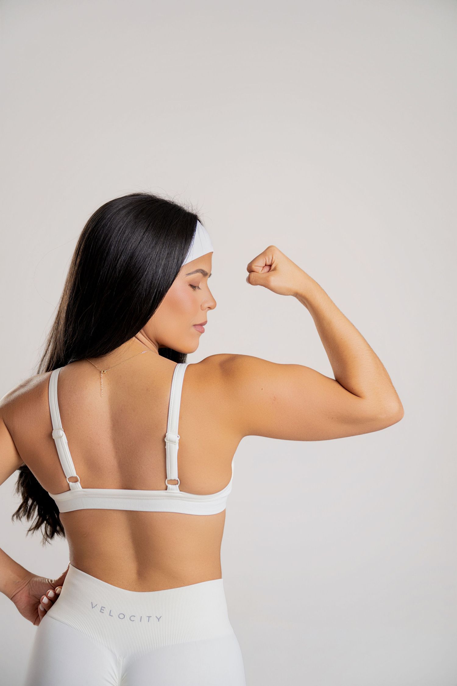 Woman in white Velocity sports bra and leggings flexing bicep, showcasing athletic activewear from behind.