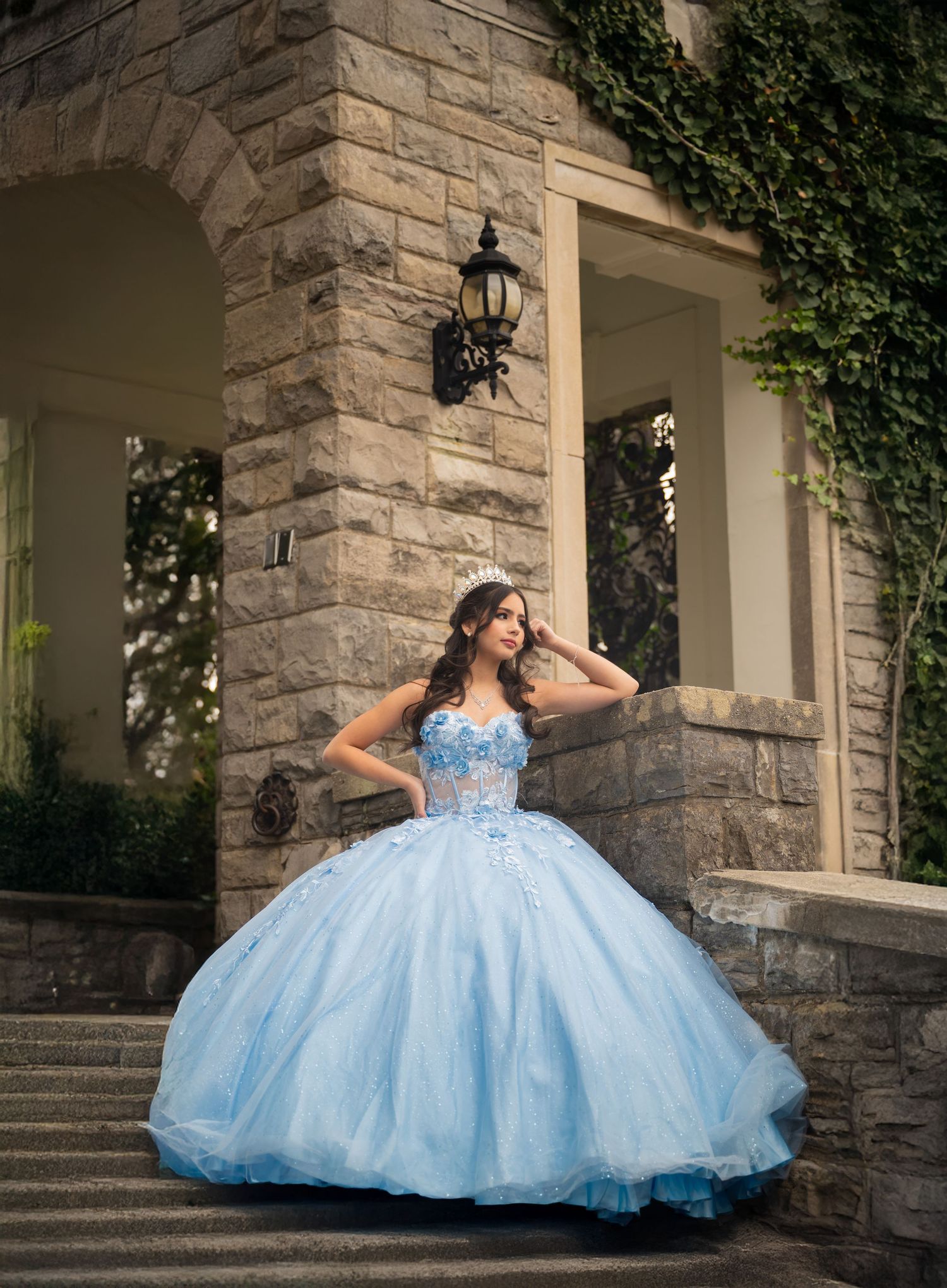 Young woman in a light blue ball gown and tiara poses on stone steps outside an elegant mansion.