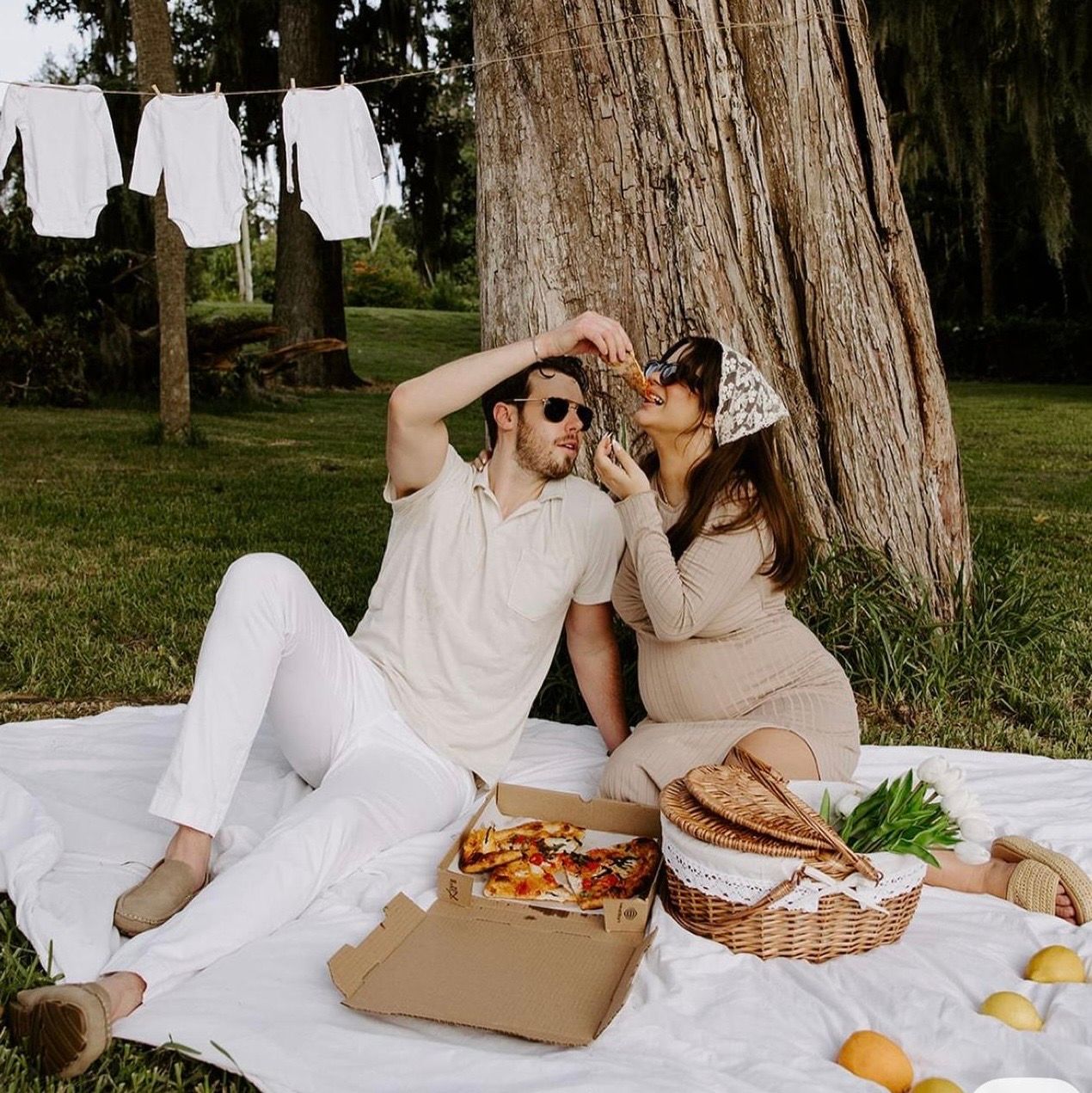 Couple enjoying a romantic outdoor picnic under a large tree with pizza, basket, and baby clothes hanging nearby.