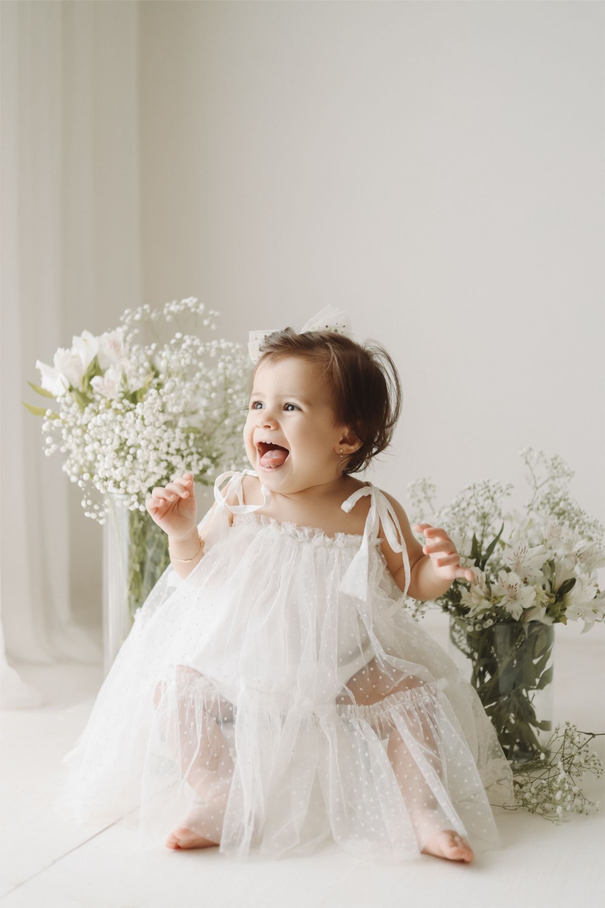 Smiling baby girl in white tulle dress sitting among white flower arrangements in a bright studio.