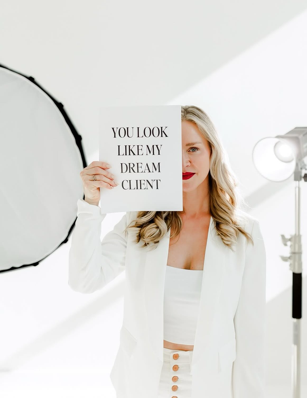 A blonde woman in a white blazer holds a sign reading You Look Like My Dream Client in a bright photography studio.