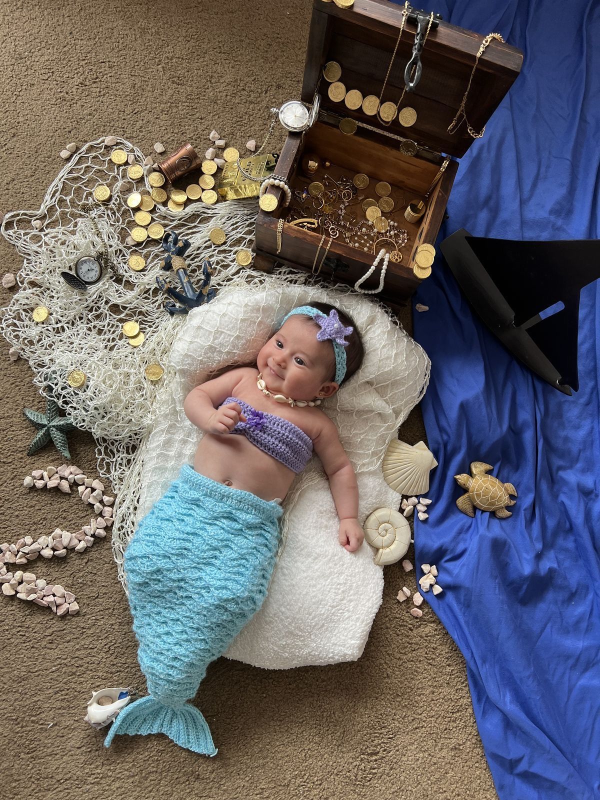 Newborn baby dressed in a mermaid costume with treasure chest, gold coins, and fishing net backdrop.