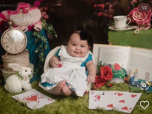 Baby girl in Alice in Wonderland costume surrounded by props including white rabbit, playing cards, roses, clock, and open book.