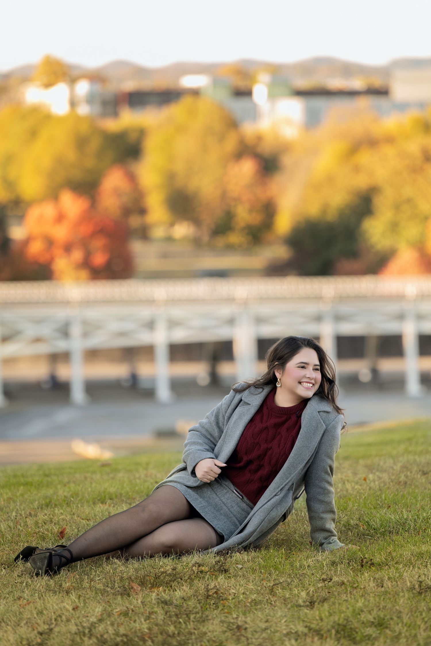 Smiling woman in a gray coat, burgundy sweater and tights sitting on a grassy hill with colorful autumn trees and a white bridge behind her.