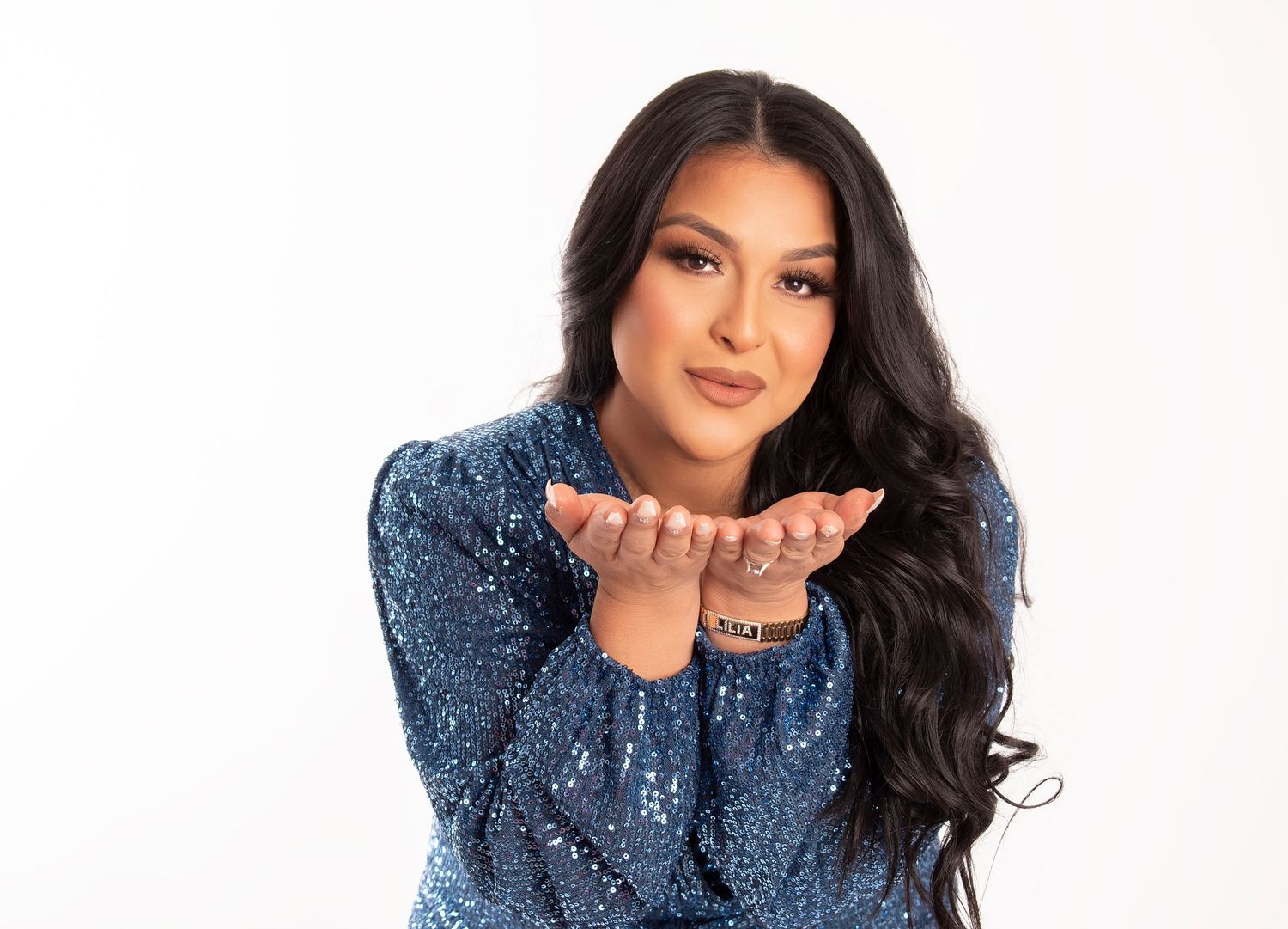 Woman with long dark wavy hair in a blue sequin top smiles and holds her cupped hands out as if blowing a kiss against a white background.