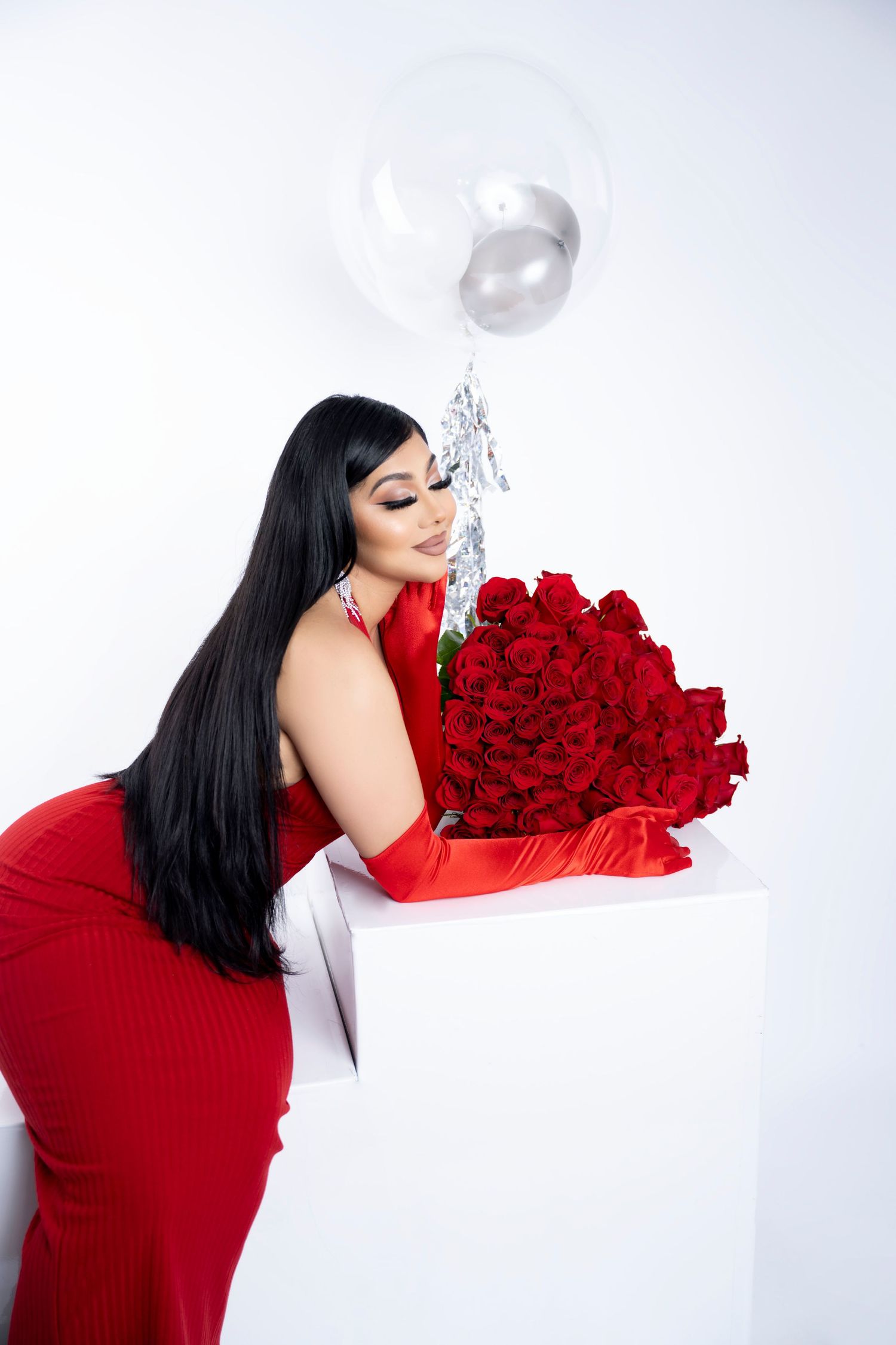 Woman in a red dress and long red gloves leans over a white pedestal holding a large bouquet of red roses beside a silver balloon.