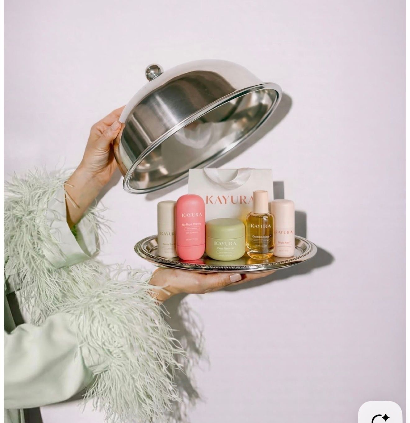 Person holding a silver cloche tray revealing Kaylie beauty skincare products in mini bottles and oils.