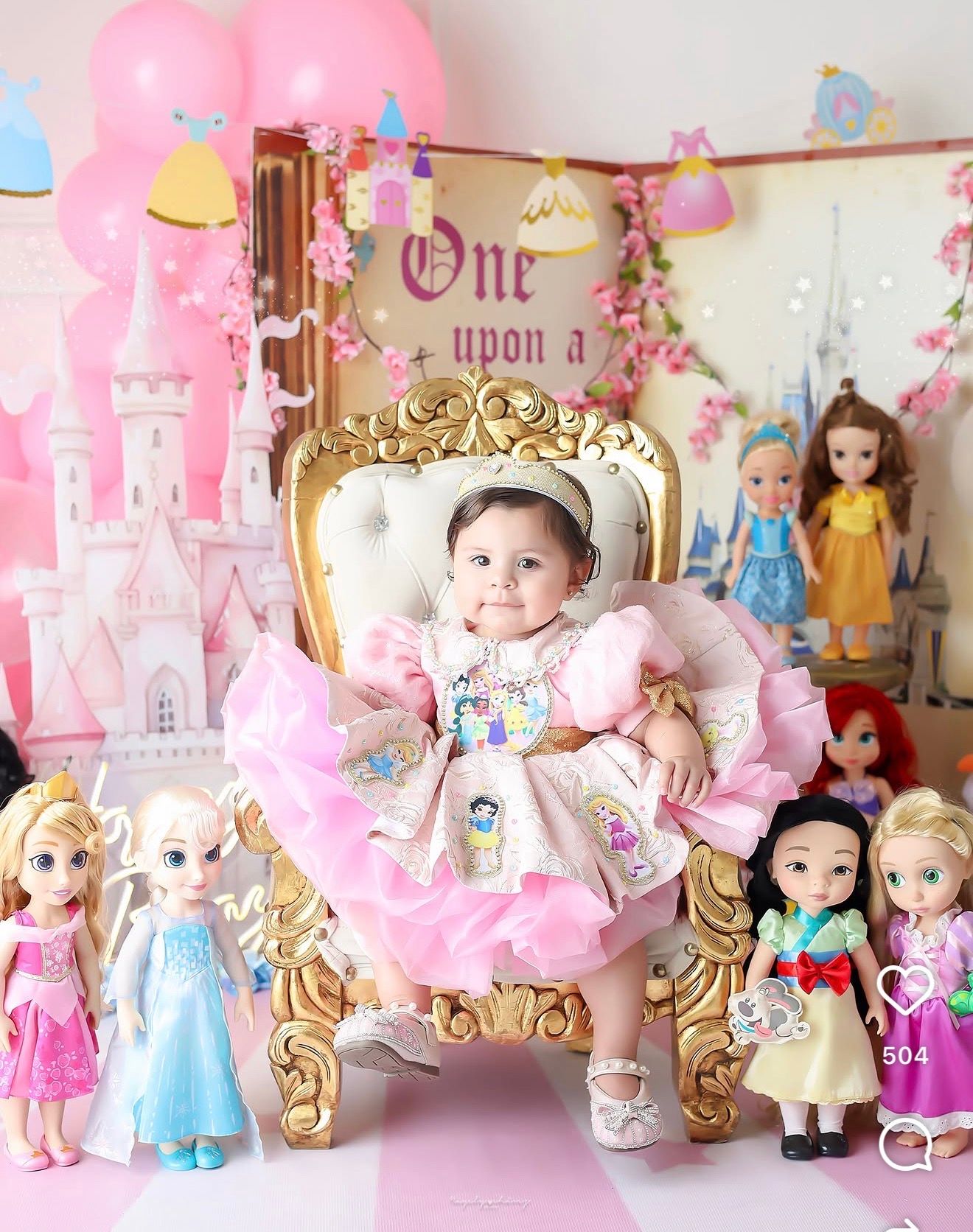 Baby girl in pink dress sits on gold throne surrounded by Disney princess dolls at a fairy tale first birthday party.