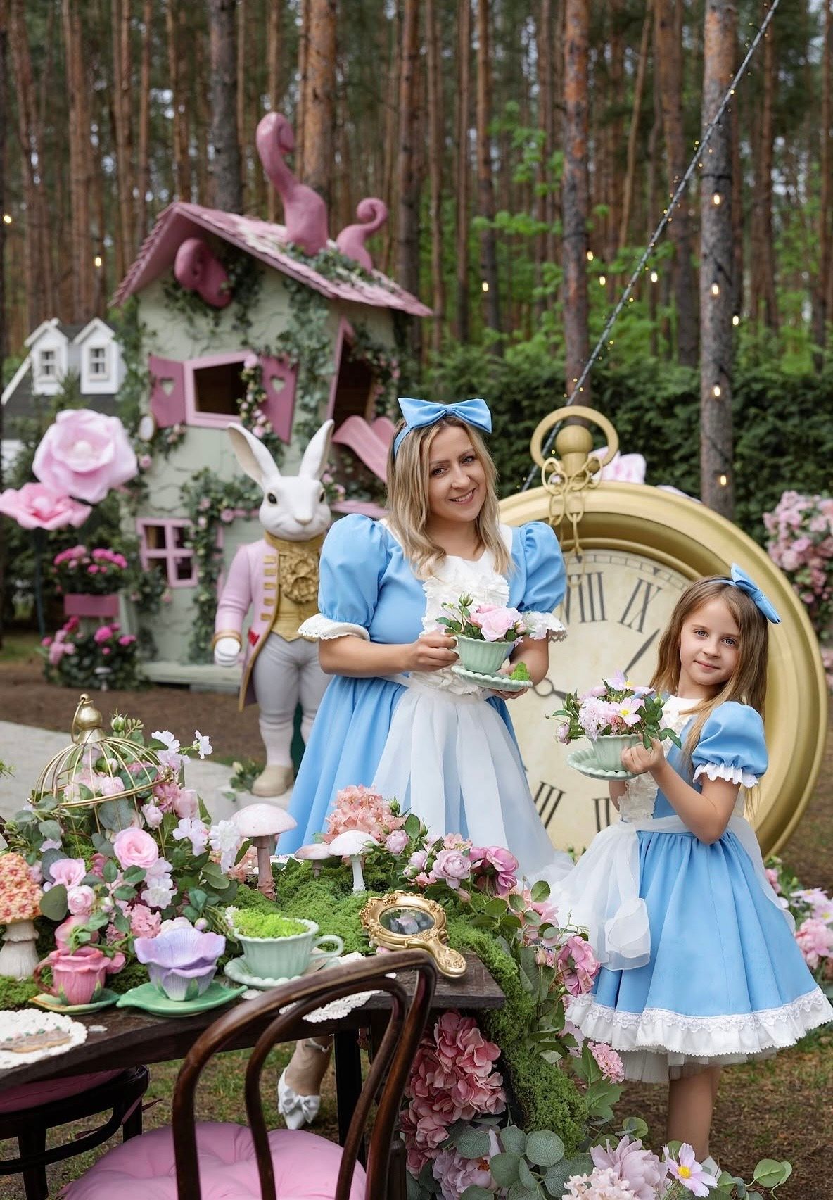 Woman and girl in Alice in Wonderland costumes at an enchanted tea party with giant clock and floral decorations.