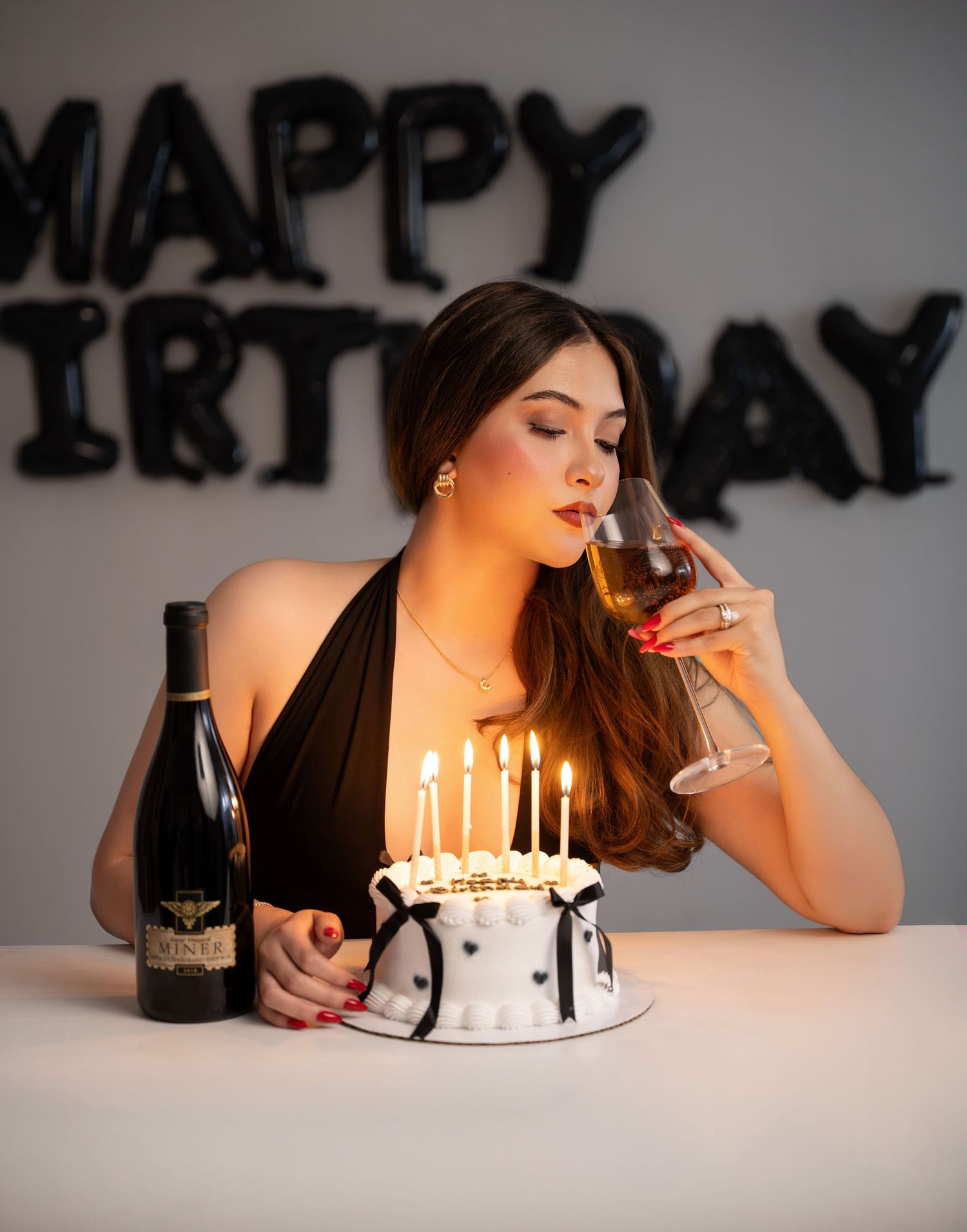 Woman in a black halter dress sips wine beside a lit black-and-white birthday cake, with black Happy Birthday balloons behind her.