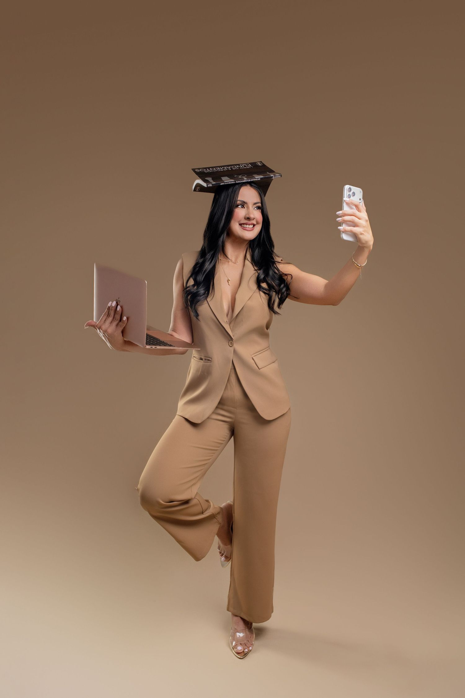 Woman in beige suit balancing book on head, holding laptop and taking selfie on brown background.